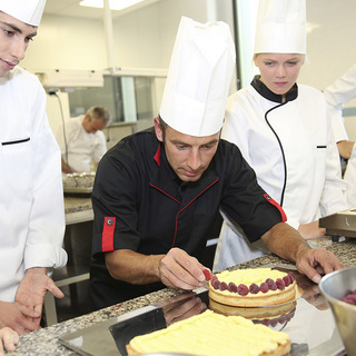 Four student chefs watching a teacher decorate a cake